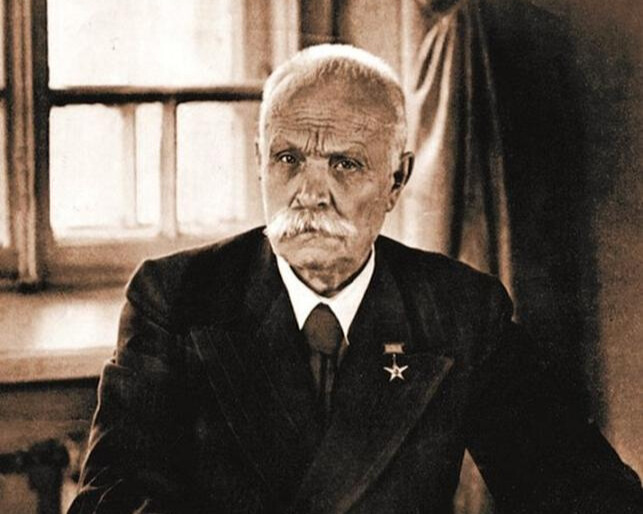 чорно-білий портрет Євгена Патона за столом в кабінеті, в темному костюмі, білій сорочці з темною краваткою, з значком у формі зірки на лацкані