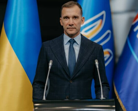 Андрій Шевченко в офіційному костюмі на фоні українського прапора та герба під час урочистої події